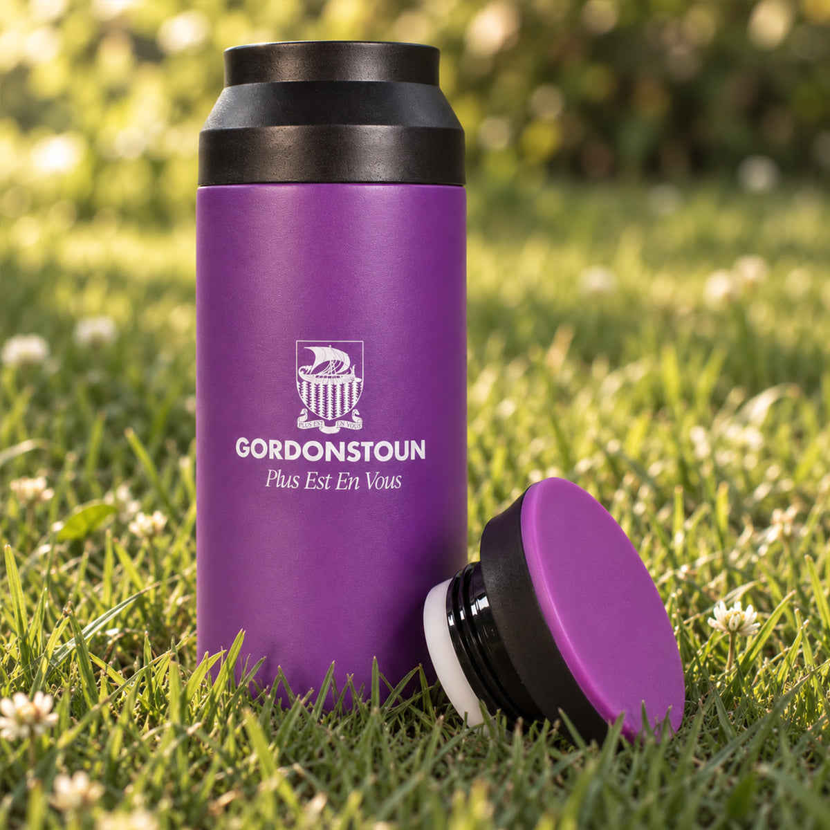 A purple insulated bottle with a black lid stands on green grass outdoors, with the matching purple lid placed beside it. The bottle has a white printed Gordonstoun logo and text on the front.