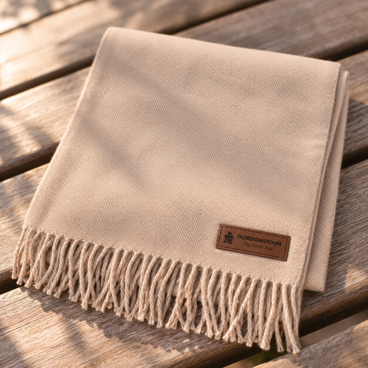 A folded beige woven scarf with fringe and a brown GORDONSTOWN label, placed on a wooden slatted surface in warm natural light.